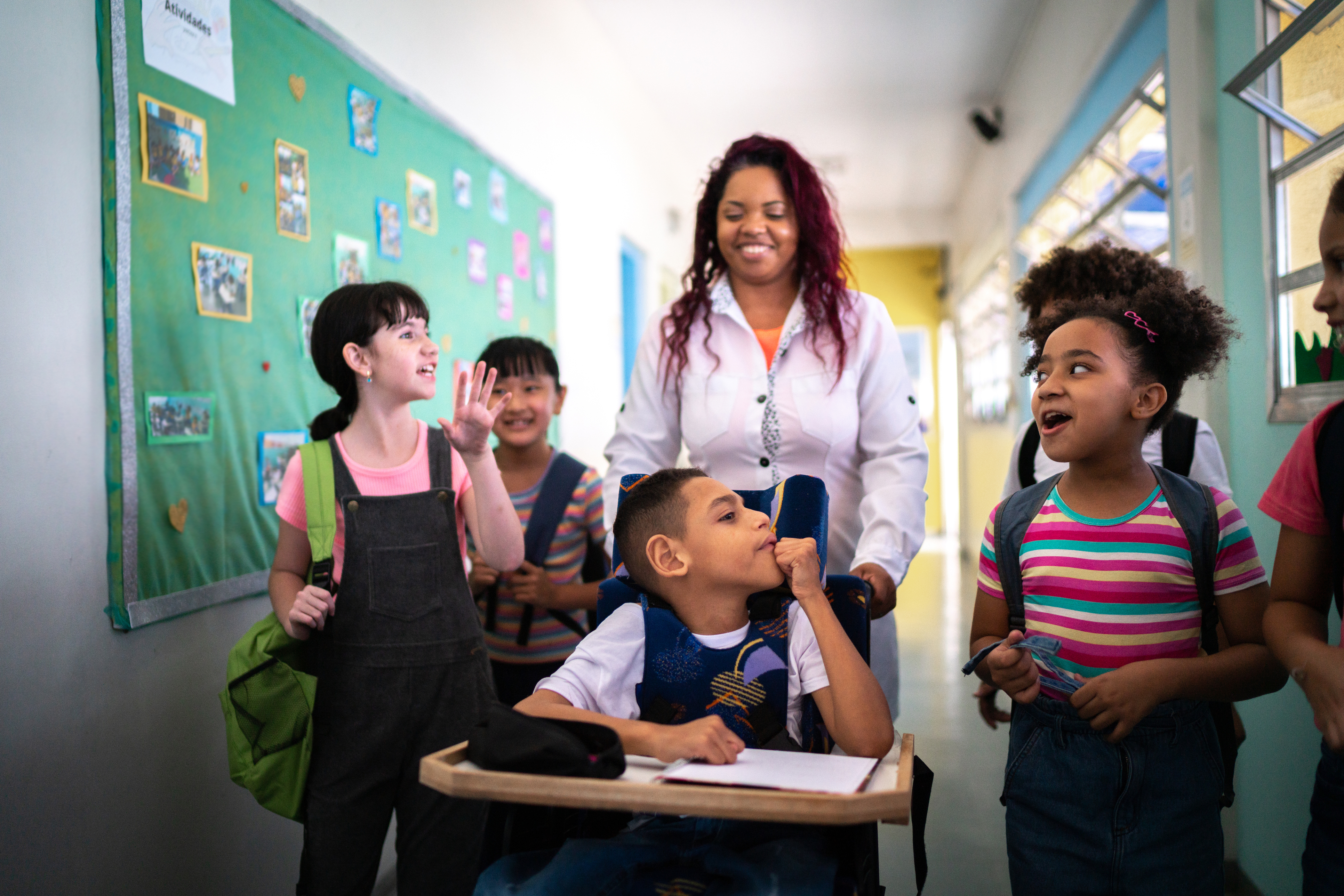 A group of six elementary-aged students in mid-conversation make their way down a school hallway with their teacher. The student in the centre of the group uses a wheelchair, which is being pushed by the teacher. The rest of the students cluster around their peer’s wheelchair, smiling and gesturing with their hands as they talk. 