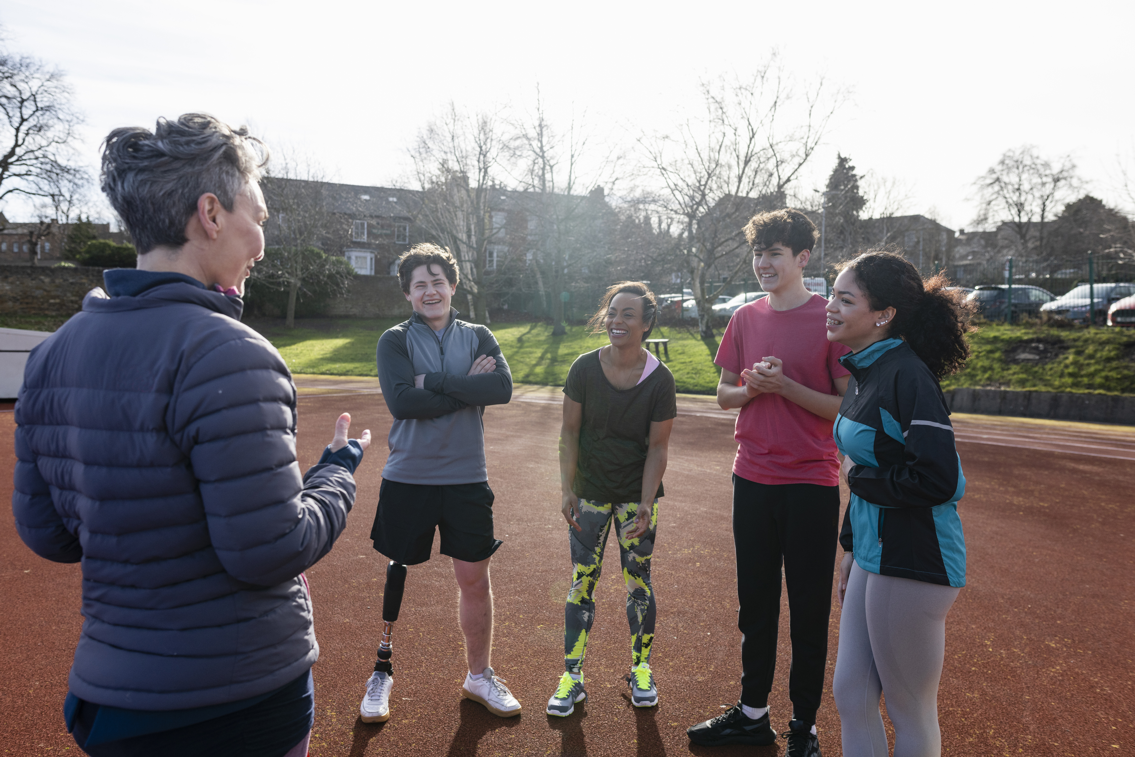 quatre élèves se tiennent face à leur professeur sur une piste extérieure à la fin de l'automne. Le professeur, qui a des cheveux gris et courts et porte une veste grise, a le dos tourné et fait des gestes avec ses mains. Tous les élèves portent des vêtements de sport et présentent une grande variété de teintes de peau, d'expressions de genre et de textures de cheveux. L'un des élèves est amputé et porte une jambe prothétique.