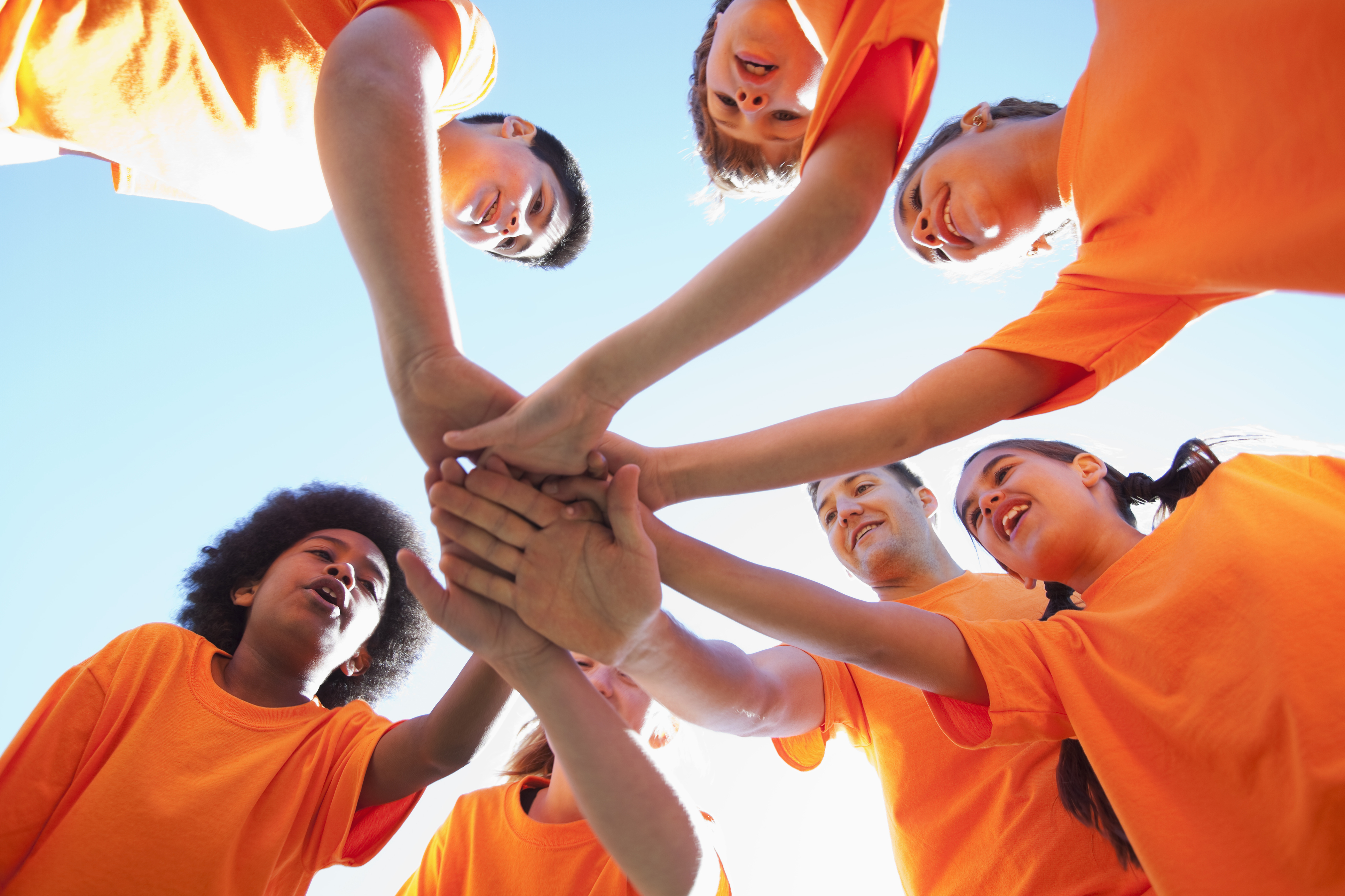 A low-angle image of six youth and their coach, wearing matching orange t-shirts, standing in a circle and stretching their hands towards the middle. The camera is positioned under the pile of hands. The youth display a variety of skin tones, hair colours and textures, and gender presentations.  