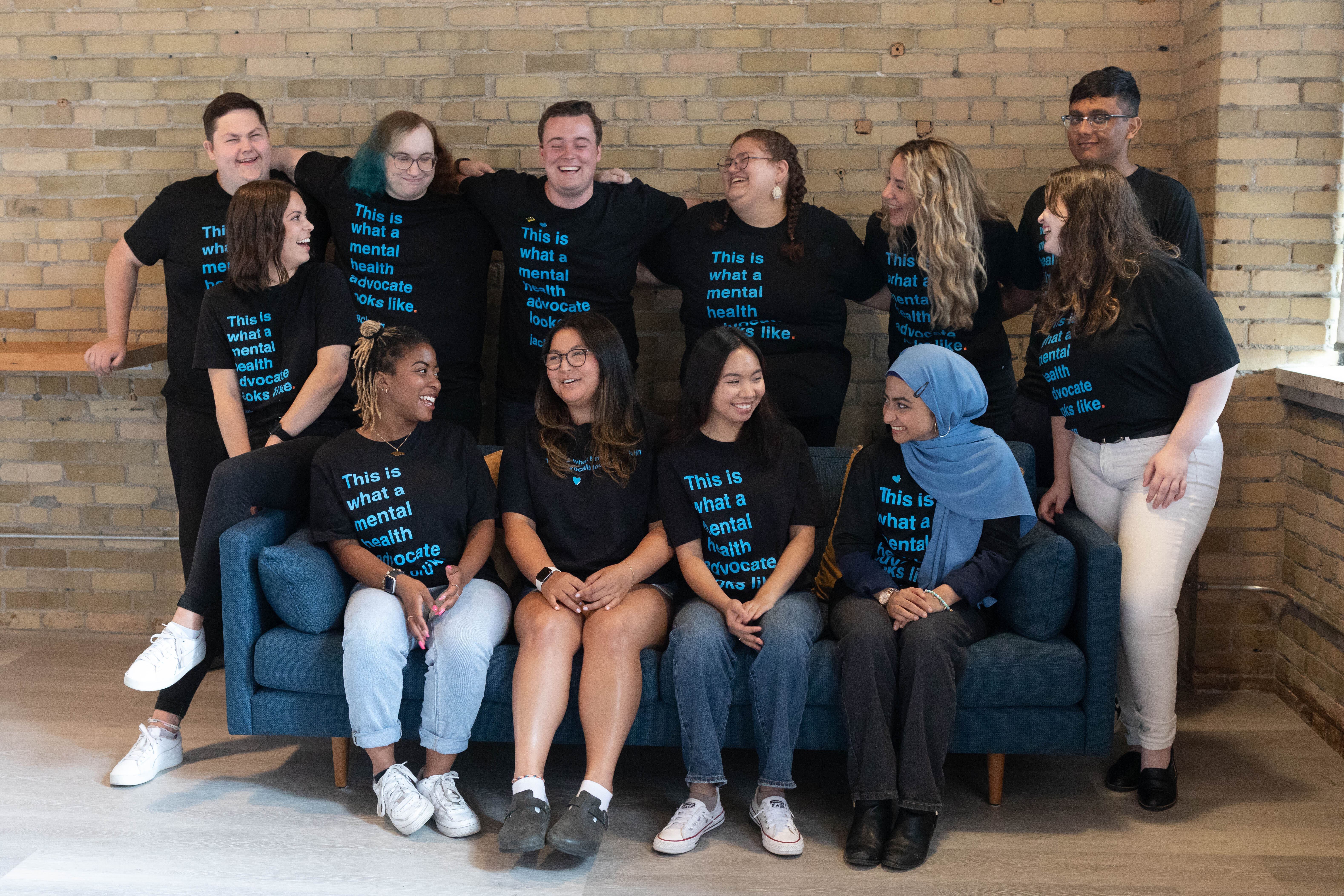 A group of young people sit on or stand around a blue couch against a plain brick wall. The group is diverse and displays a wide range of skin colour, gender expression, and cultural background. All of the youth wear the same Jack.org black T-shirt with blue text that reads: “This is what a mental health advocate looks like.” The youth are smiling with their arms around each other and laughing.
