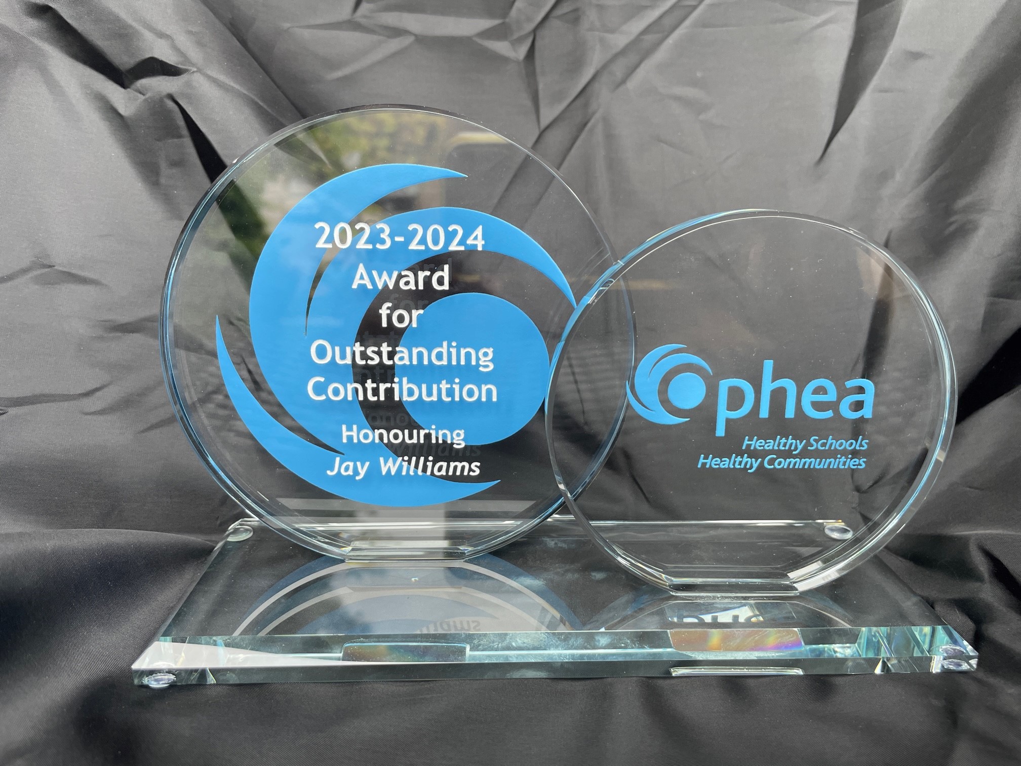 The 2023-2024 Ophea Award for Outstanding Contribution is a clear glass object composed of two circles. The circle on the right bears the text “2023-2024 Award for Outstanding Contribution Honouring Jay Williams” superimposed over Ophea’s “O” logo. The circle on the left is slightly smaller and bears Ophea’s full logo with the tagline “Healthy Schools, Healthy Communities”. 
