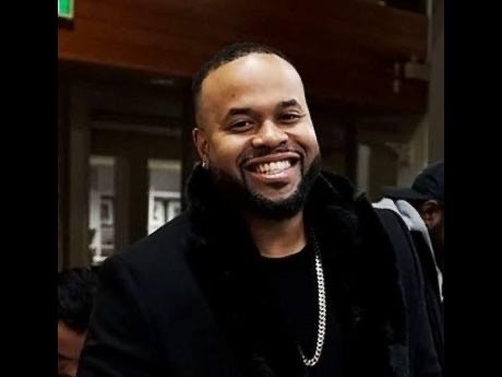 Jay Williams, a Black man with short dark hair wearing dark clothes and a silver chain, smiles warmly at the camera. 