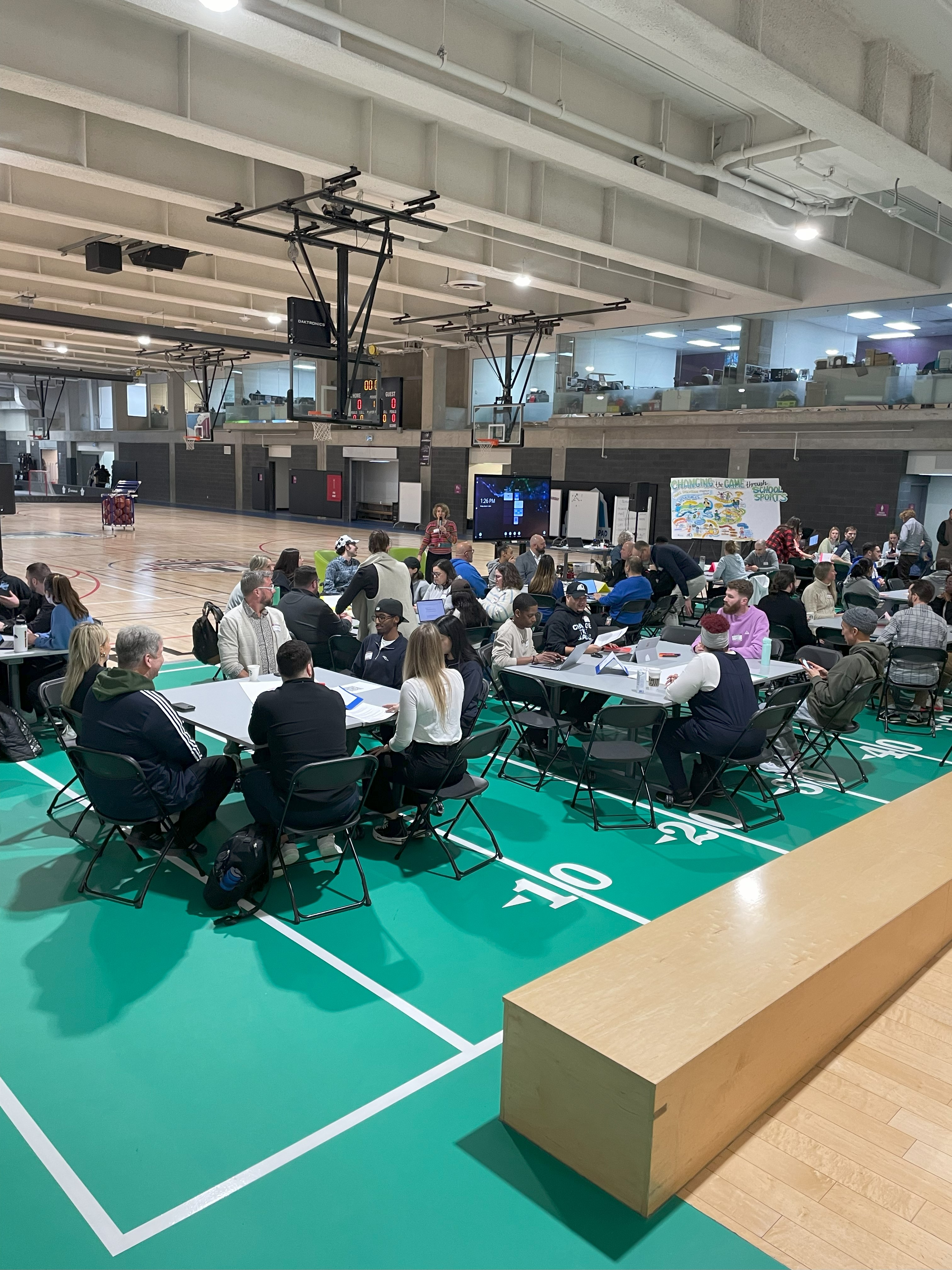 Un grand groupe de personnes est assis autour de tables sur un terrain de basket au MLSE LaunchPad. À l'avant du groupe, une femme blanche aux cheveux blonds bouclés parle dans un microphone. Elle se tient devant un tableau blanc interactif.