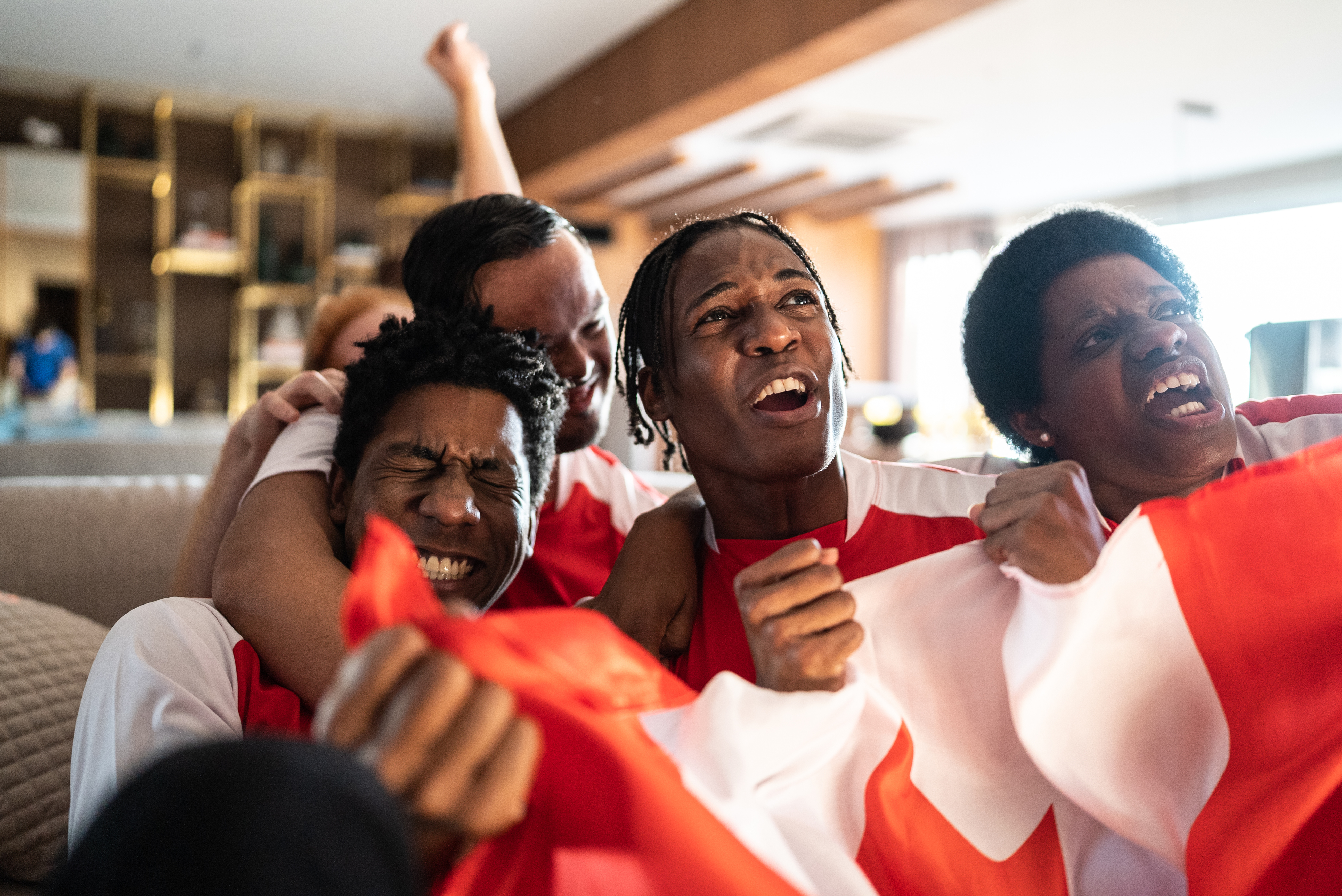 Four individuals sit clustered on a sofa watching and enthusiastically cheering on a sporting event on the television. The three Black people sitting in front of the couch clutch a Canadian flag, and everyone is wearing matching red and white. 