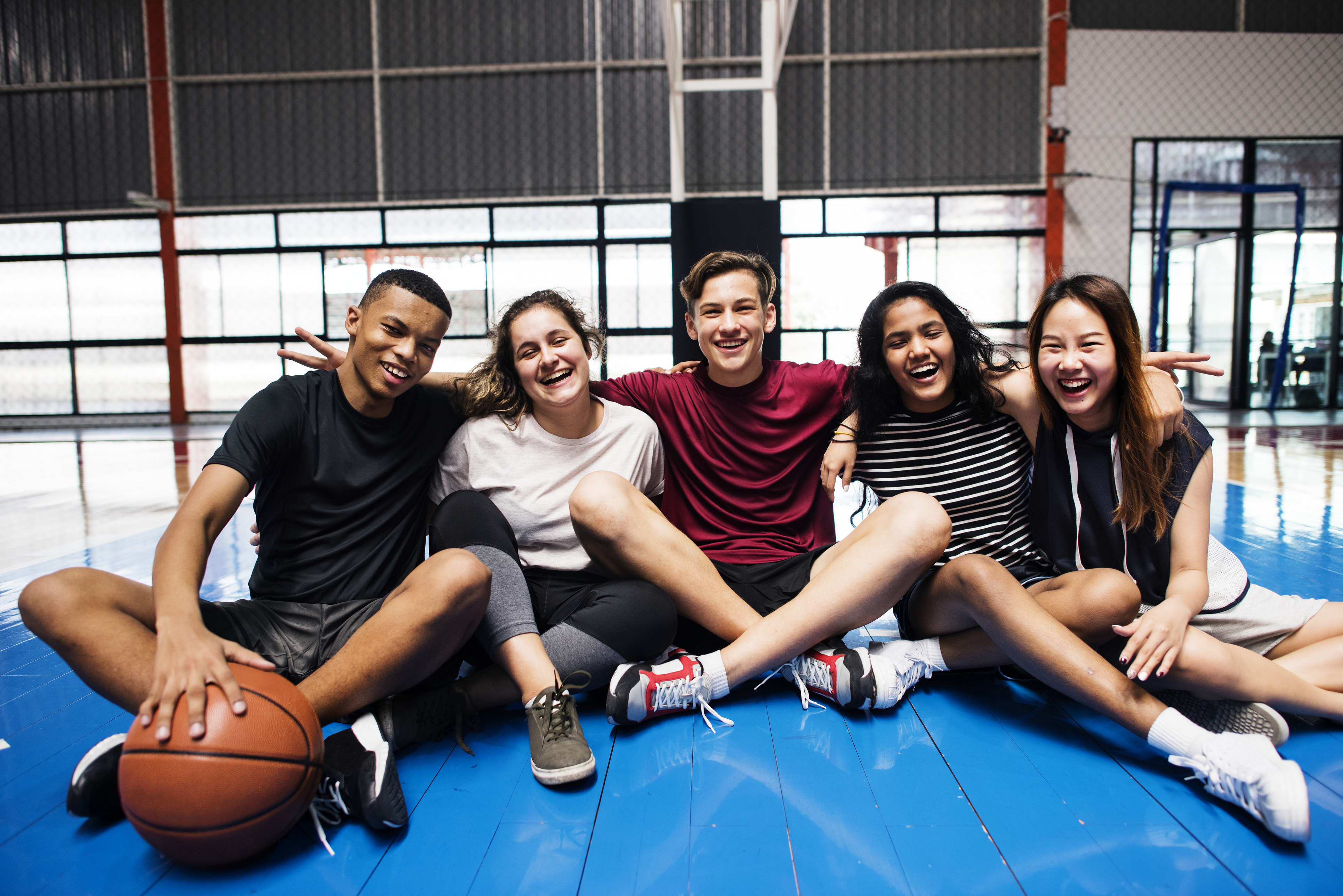 Five smiling students sit clustered together on the floor of a school gymnasium, one holding a basketball. They are dressed in athletic clothes. 