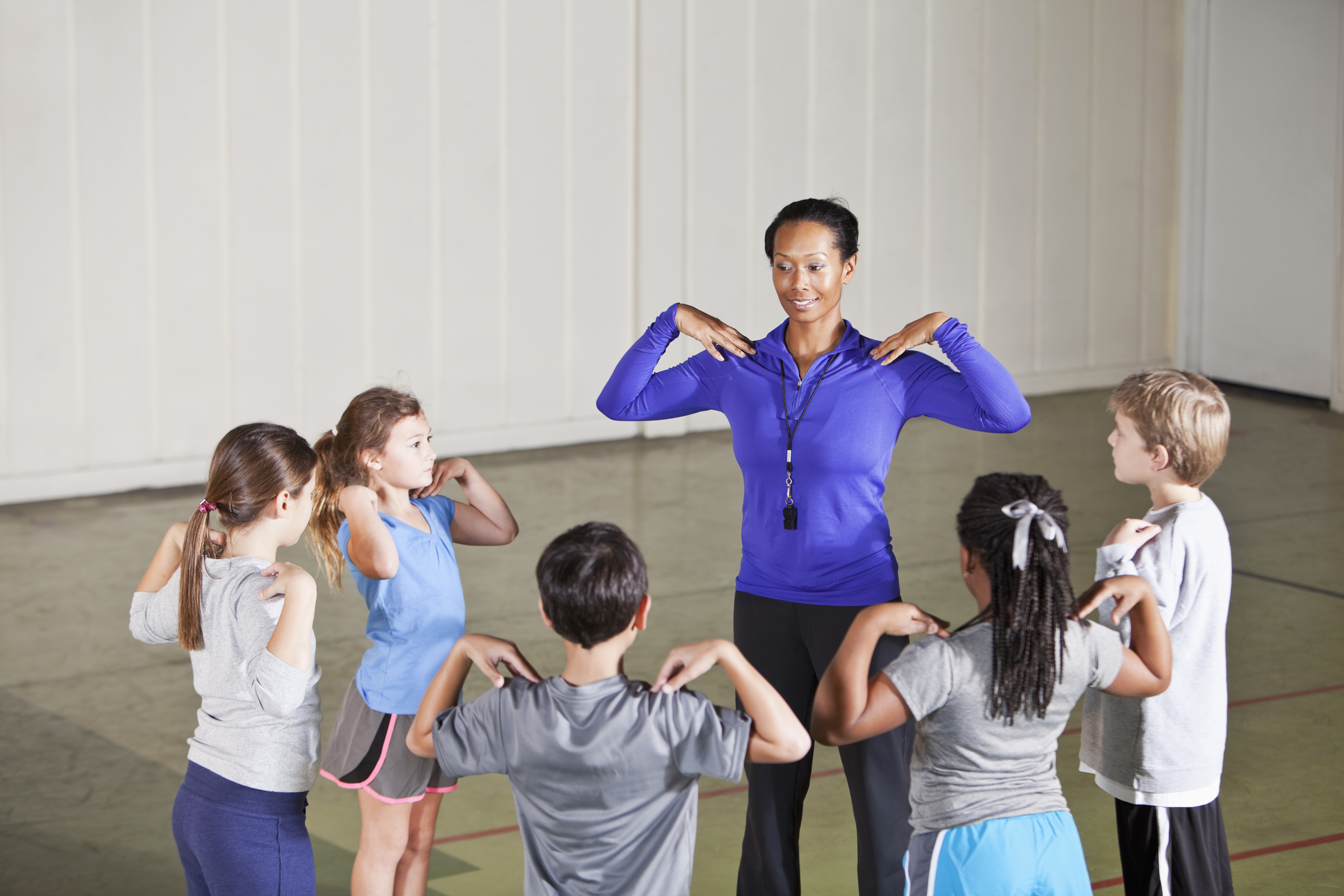 A physical education educator leads an exercise session with five children, who mimic her movements, in a school gymnasium. They all have their hands on their shoulders. 