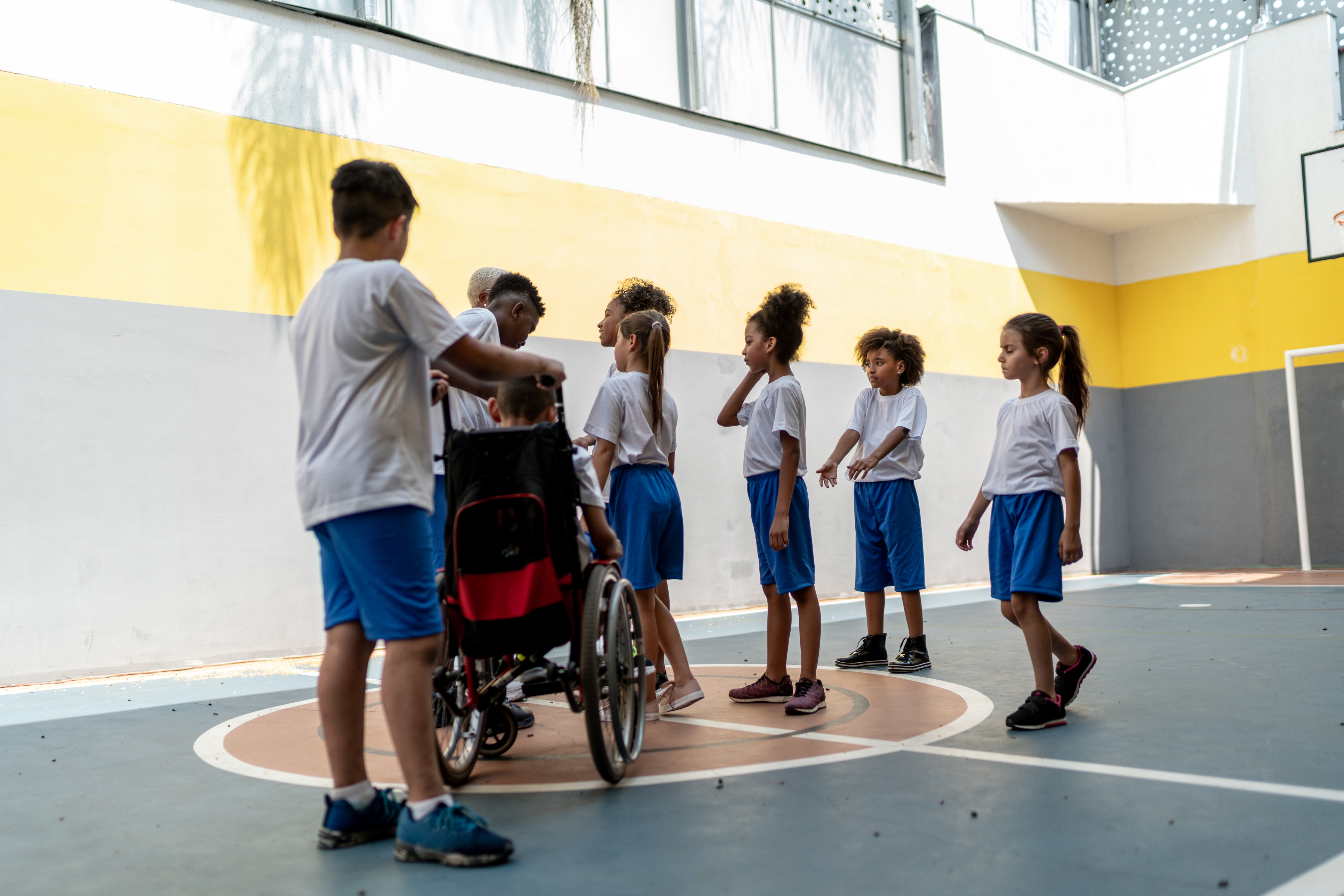 In a gymnasium, a diverse group of students wearing matching uniforms cluster around a teacher for instruction. The students display a range of gender presentations, skin tones, and hair textures. One of the students uses a wheelchair and is being pushed by one of their peers to join the rest of the group. 