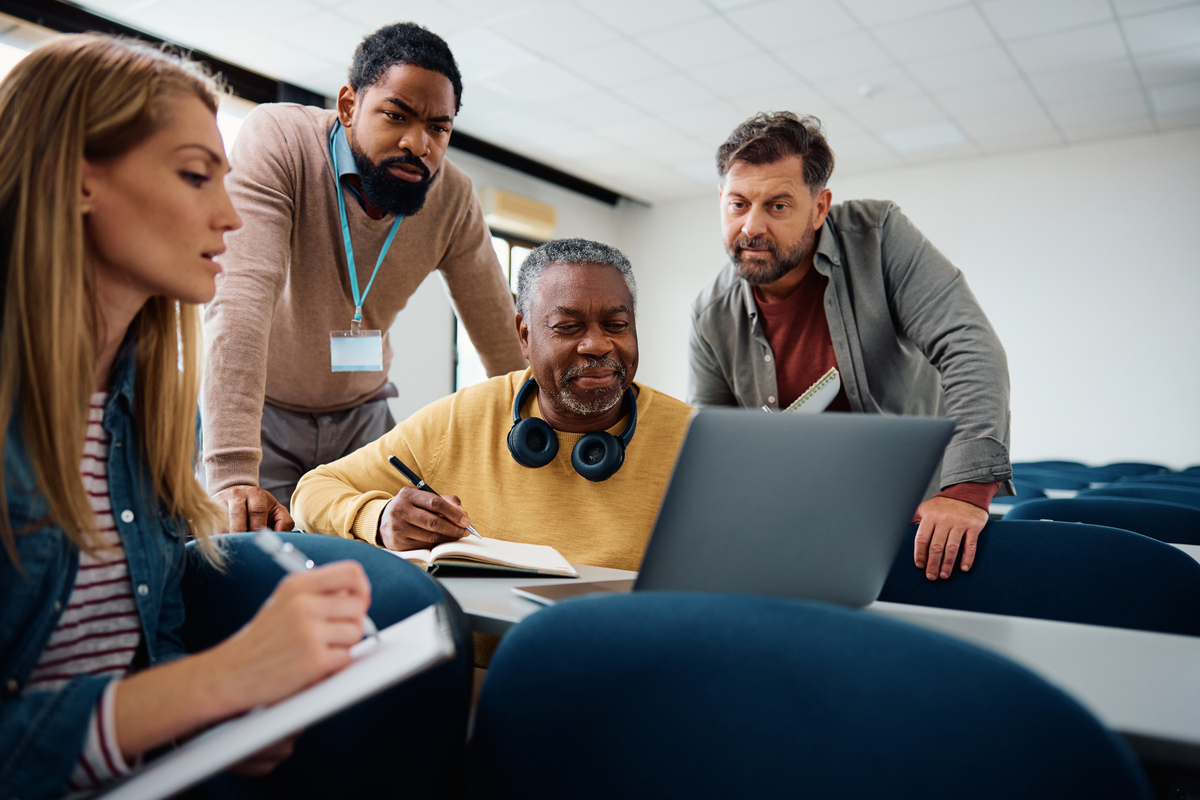 Four diverse individuals gather around a laptop in a classroom. One is writing notes, while others watch intently, conveying collaboration and focus.