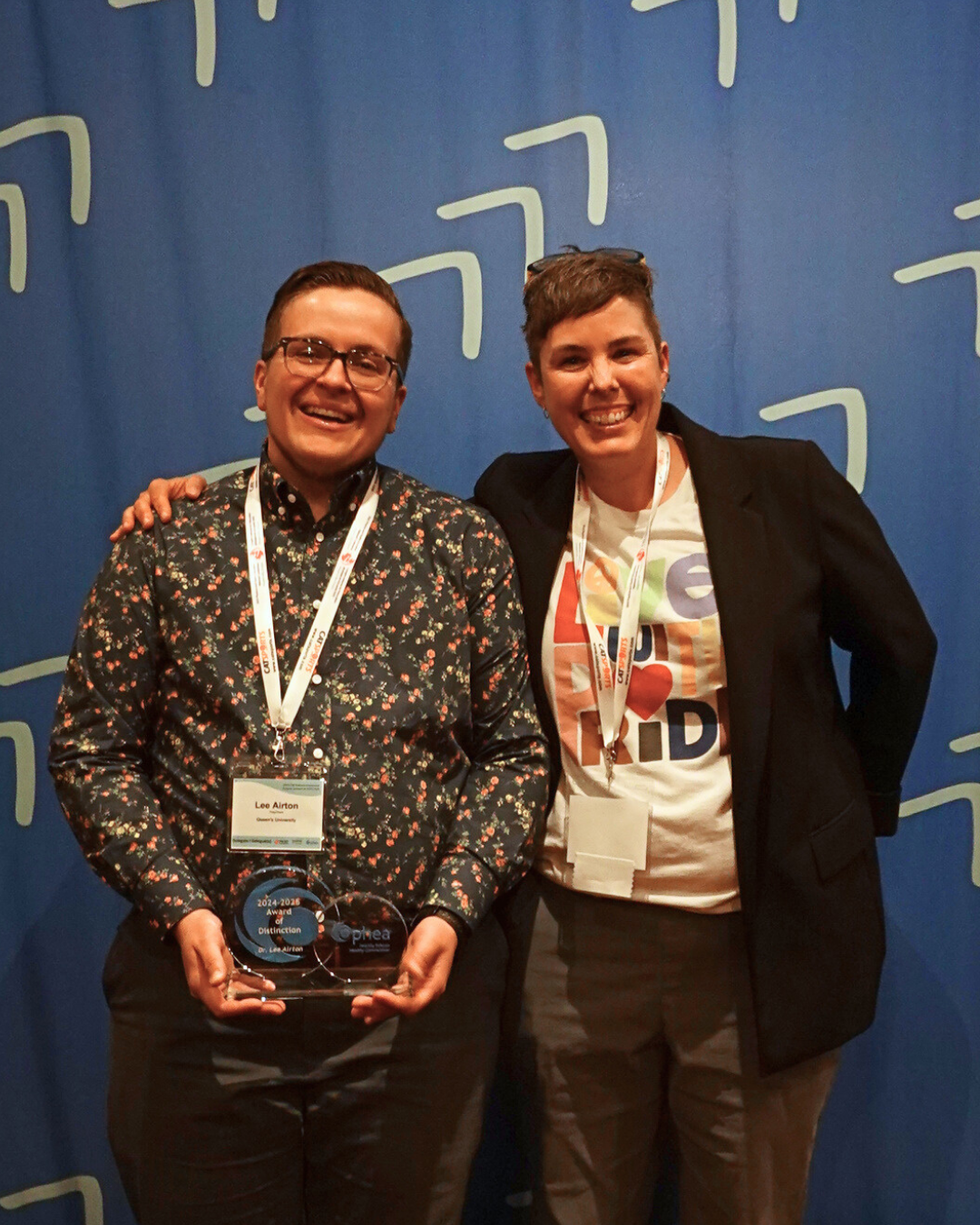 Two smiling individuals stand side by side. Both are smiling and one is holding the “Ophea Award of Distinction”