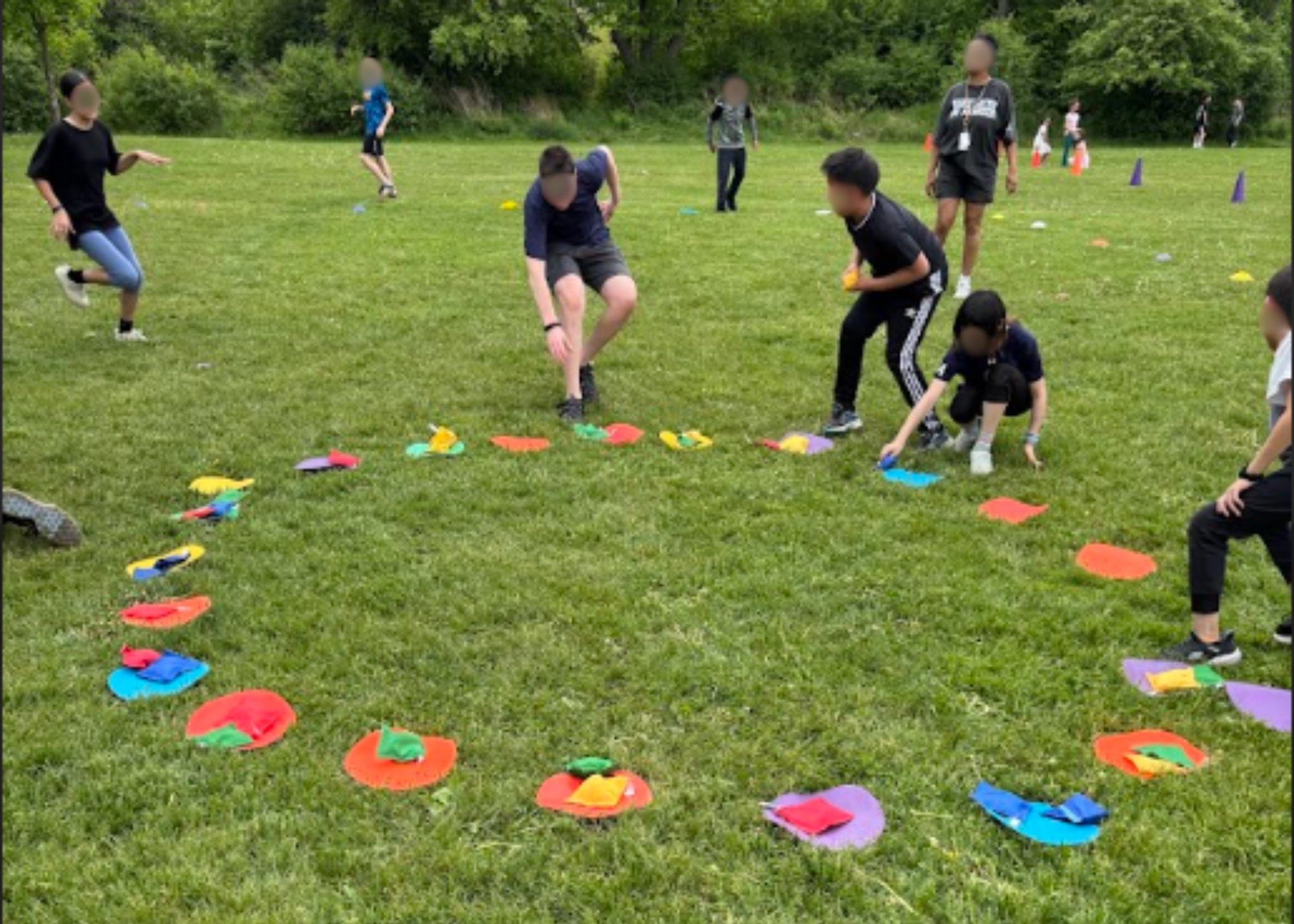 Des enfants sont rassemblés à l'extérieur pour jouer à un jeu avec des sacs de fèves multicolores disposés en cercle sur l'herbe. L'atmosphère est active et vivante.