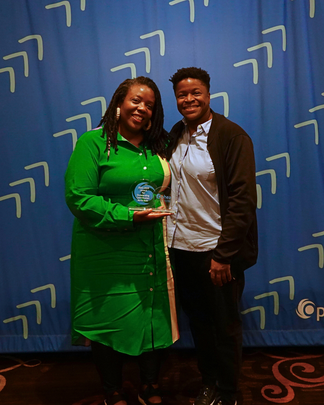 Two people smile as they pose with the “Ophea Award for Outstanding Contribution” against a blue backdrop with a geometric pattern.