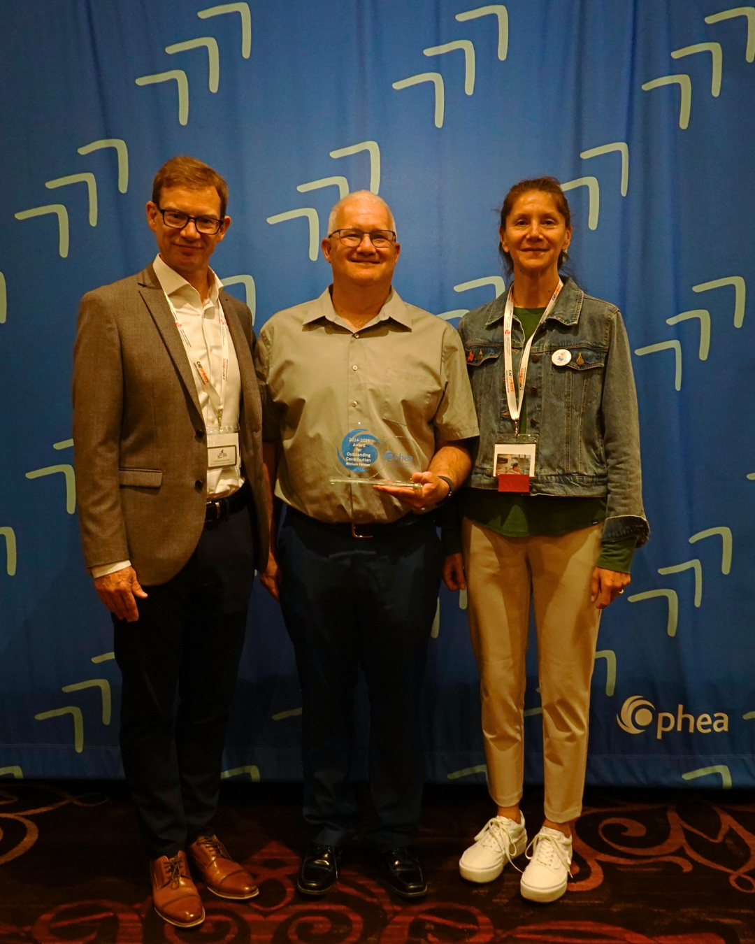 Three people stand smiling in front of a blue backdrop with a pattern. The person in the center holds the” Ophea Award of Outstanding Contribution”.