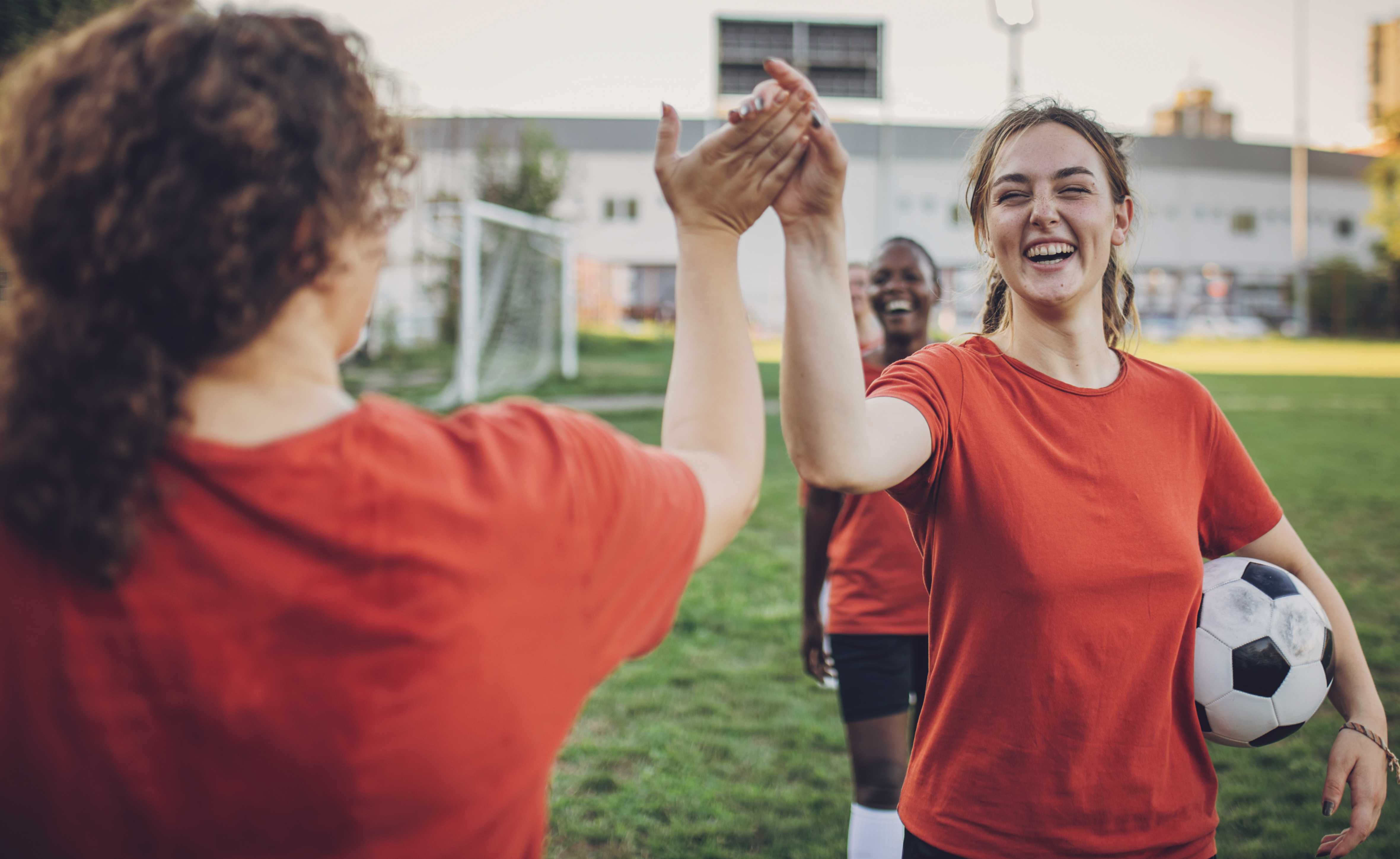 A young woman in a soccer uniform gives a high-five to her teammate on a field. 