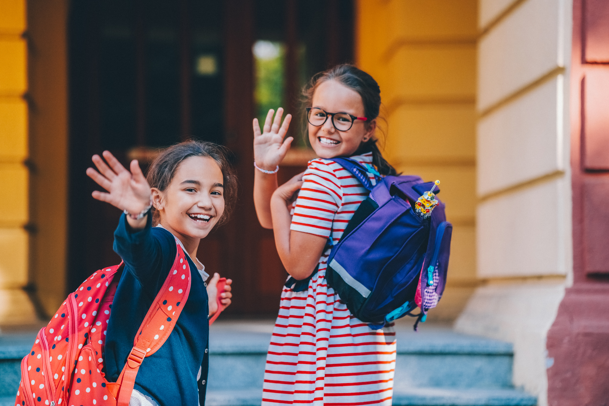 Deux filles souriantes avec des sacs à dos saluent joyeusement la caméra. Elles se tiennent sur les marches de l'école et portent des sacs à dos.