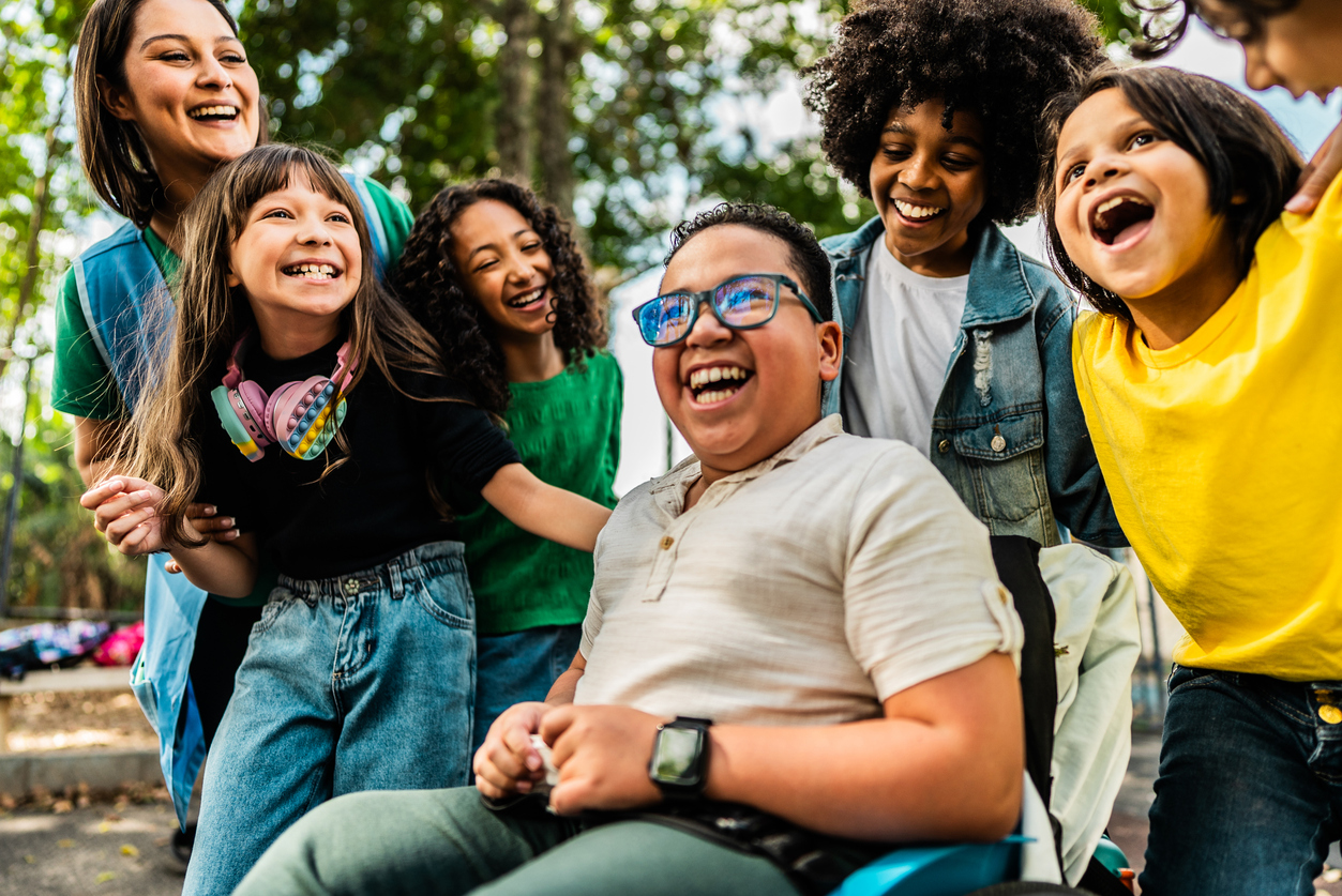 A group of children and an adult smile and laugh together outdoors. One child is seated in a wheelchair, and others stand around, appearing happy and engaged.