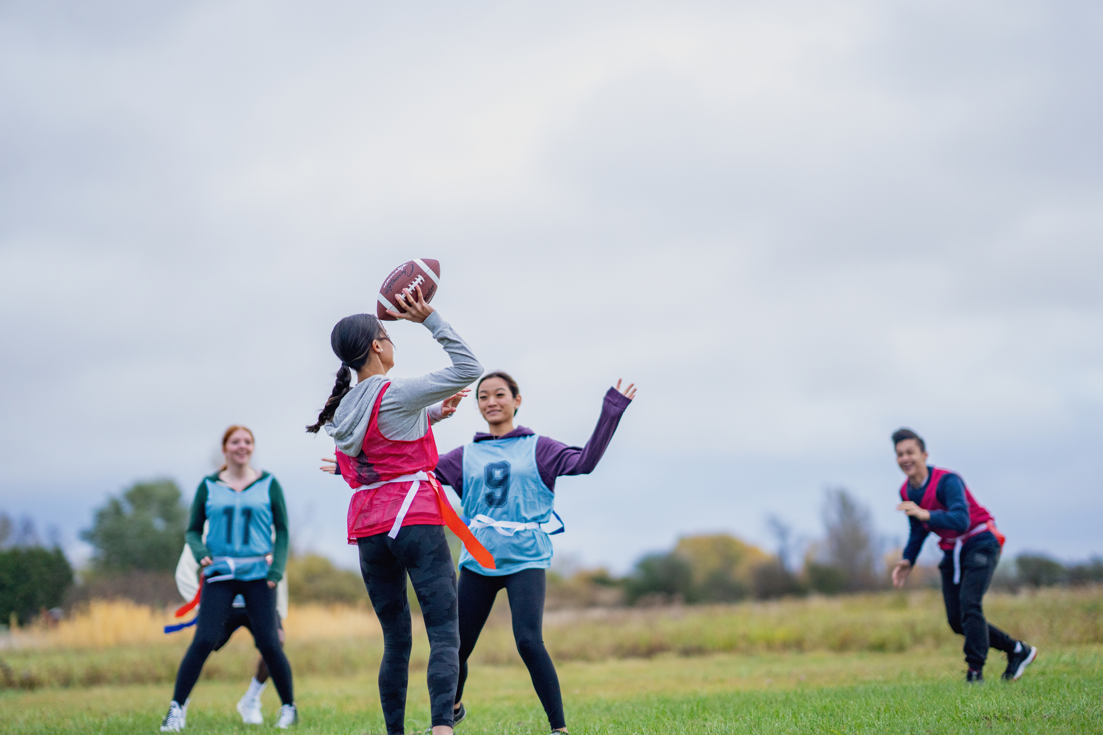 Trois filles jouent au flag football sur un terrain en herbe, l'une d'elles attrapant une passe. Elles portent des maillots colorés et semblent concentrées et joyeuses sous un ciel nuageux.