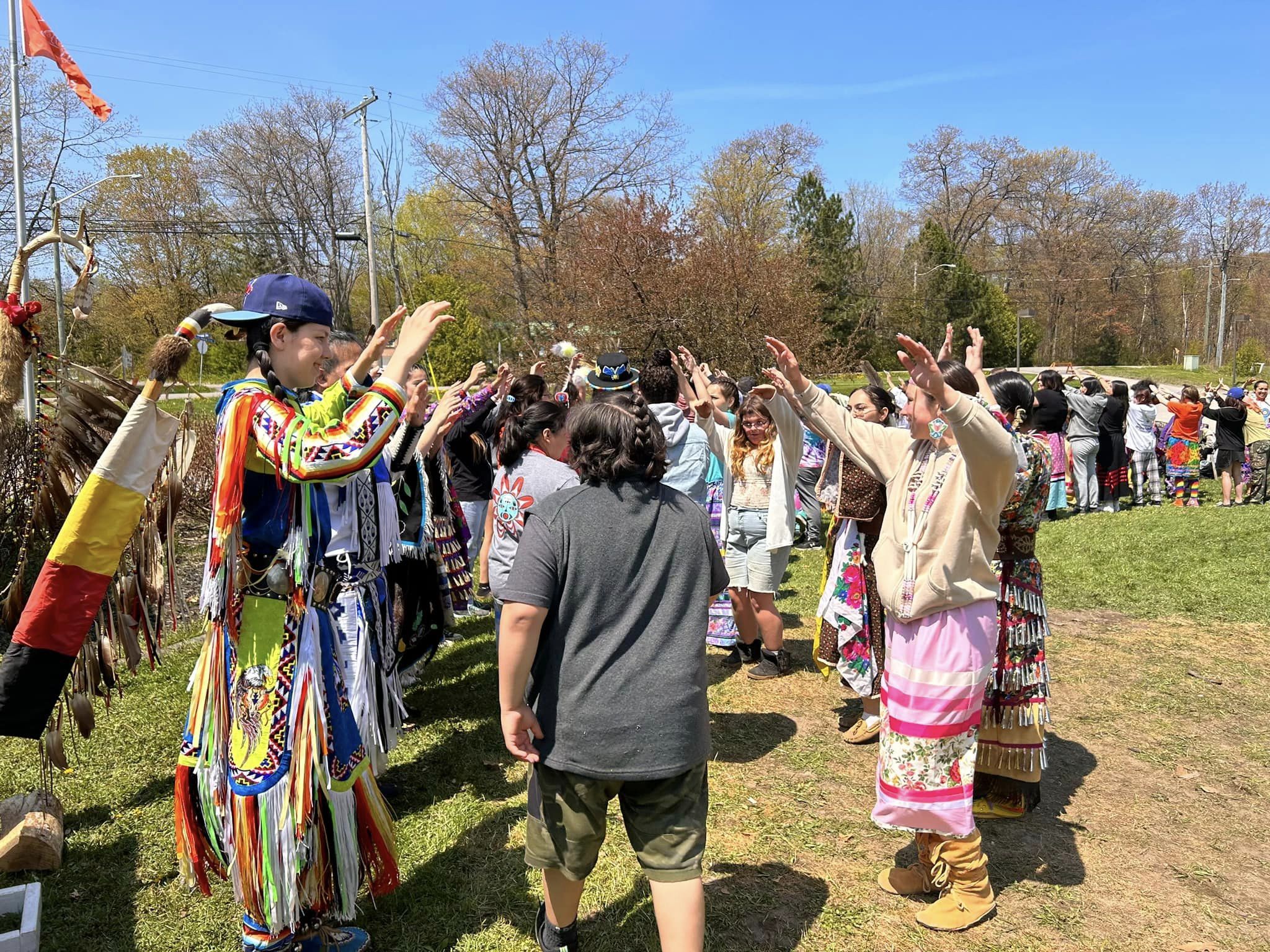 People in traditional attire dance outdoors on a sunny day. Colorful regalia with tassels and feathers create a vibrant, joyful atmosphere.