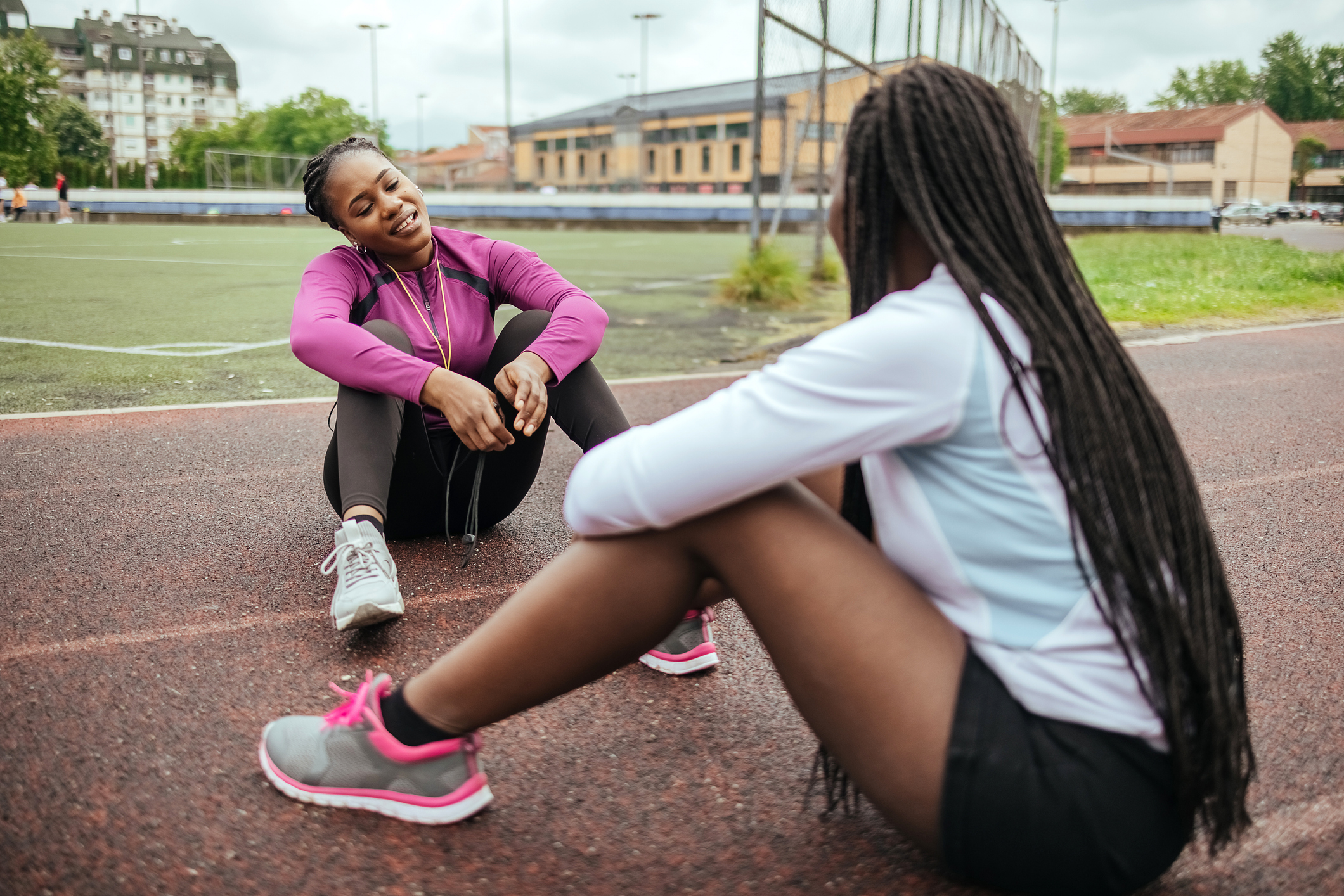 Un entraîneur et un élève athlète sont assis sur une piste de course, souriants et détendus après leur entraînement. Ils portent des vêtements de sport. Un terrain et des bâtiments sont visibles à l'arrière-plan. 