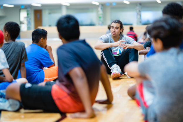 A group of youth sit on a gymnasium floor.