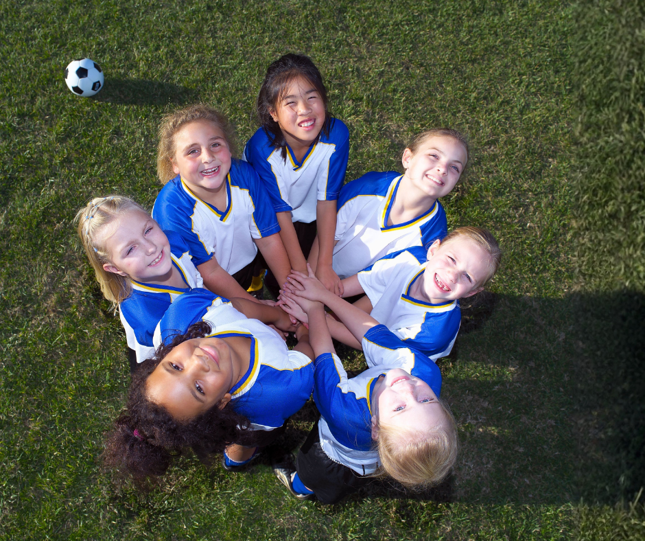 A diverse group of girls stand in a circle on a soccer field wearing team uniforms with their hands in the middle.