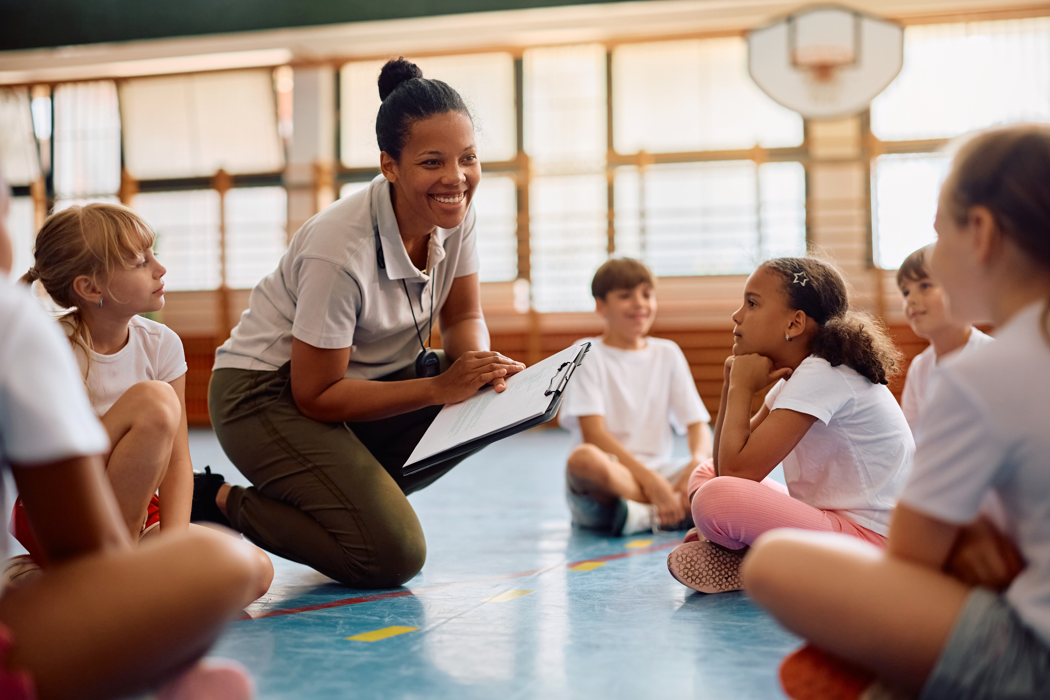 A physical education teacher and her elementary students talking on the floor of school gym.