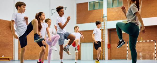 Group of six elementary students of various genders and races with their teacher inside a school gymnasium. They are raising one knee and the opposite arm to mimic the act of running.