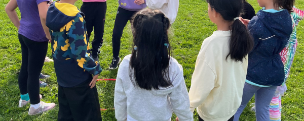 A group of students stand in a circle around a hula hoop that has been laid on the grass of a school field. Most of the students have their back to the camera and are listening to a student wearing a surgical mask. 