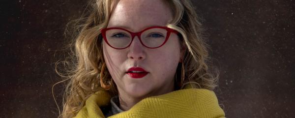 Une femme blanche aux longs cheveux blonds se tient à l'extérieur, regardant calmement la caméra. Elle porte des lunettes à monture rouge, du rouge à lèvres rouge et un manteau à chevrons jaune vif. 