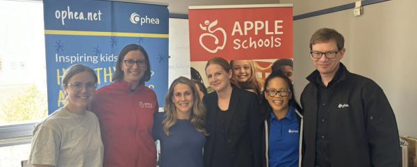 Six adult individuals are standing together in front of banners for Ophea and APPLE Schools. 