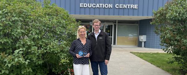 Lara Paterson, a white woman with blond hair and Ophea's 2023-2024 Award of Distinction recipient stands smiling beside Ophea Executive Director Chris Markham, a white man with glasses and brown hair. They stand in front of the Education Centre at the Limestone District School Board. 