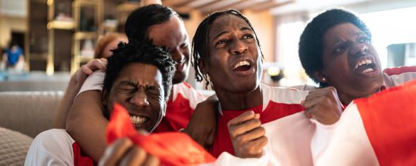 Four individuals sit clustered on a sofa watching and enthusiastically cheering on a sporting event on the television. The three Black people sitting in front of the couch clutch a Canadian flag, and everyone is wearing matching red and white. 