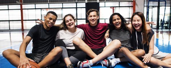 Five smiling students sit clustered together on the floor of a school gymnasium, one holding a basketball. They are dressed in athletic clothes. 