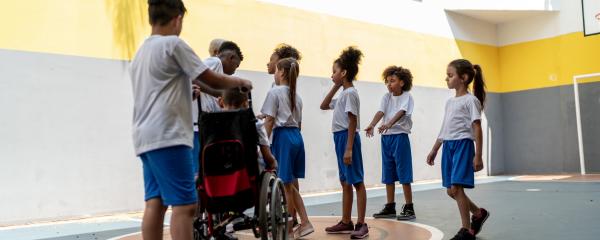 In a gymnasium, a diverse group of students wearing matching uniforms cluster around a teacher for instruction. The students display a range of gender presentations, skin tones, and hair textures. One of the students uses a wheelchair and is being pushed by one of their peers to join the rest of the group. 