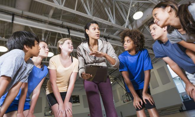 A teacher and a group of students stand in a huddle in the middle of a gymnasium. The teacher is holding a clipboard and speaking to the students, who are listening intently. Everyone in the photo is wearing athletic clothing.
