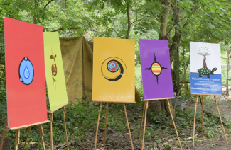 Picture of 5 paintings set up on easels in a forest: four paintings represent the elements; fire (or sun), air, water, and earth; one painting of a turtle with a tree growing out of its shell, Natural Curiosity, A Resource for Educators poster. Tent in the background. 