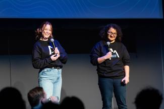 Two people on stage delivering an address at a National Jack.org Summit in support of youth mental health. One individual has white skin, long red hair tied back in a ponytail, and is wearing light blue jeans. The other has medium olive-toned skin, shoulder-length dark curly hair, and is wearing dark jeans and red nail polish. Both speakers hold microphones and are smiling at the audience.