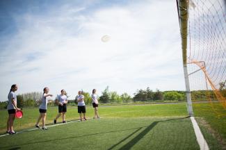 Five students wearing athletic uniforms stand on a turf field in front of a soccer net. One of the students has just thrown a white frisbee, which the students are watching fly towards the net. 