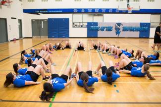 A large group of students lie on their backs, forming a large circle and performing an exercise with their legs pointing inwards on a gymnasium floor. The students wear matching athletic uniforms with blue shirts, and most have their long hair tied back in ponytails.  