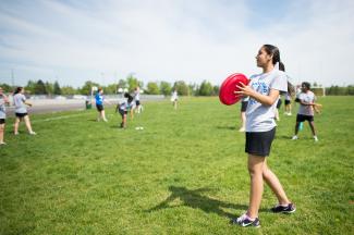 A student on an outdoor field wearing their long dark hair tied back in a ponytail and an athletic uniform prepares to throw a red frisbee to someone standing out of frame. A large group of students are visible behind them, throwing frisbees in groups and pairs. 