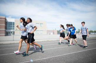 Students run and walk at various paces on a paved outdoor track. The students wear matching athletic uniform and appear to be running in front of a school yard. 