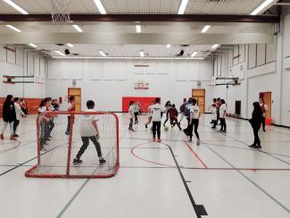A view from behind a net of a large group of students playing soccer in a gymnasium. 