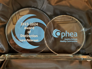 Ophea Award of Distinction 2023-2024: Lara Paterson. The Ophea Award is a clear glass object with Ophea’s logo and tagline in blue: Healthy Schools, Healthy Communities. 