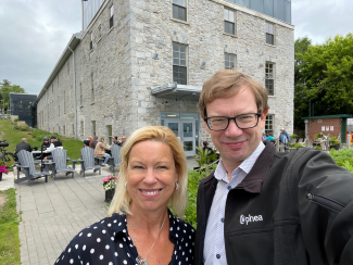 Lara Paterson, a white woman with blond hair and Ophea's 2023-2024 Award of Distinction recipient stands smiling for a selfie beside Ophea Executive Director Chris Markham, a white man with glasses and brown hair. They stand outdoors in a building courtyard with others milling in the background. 